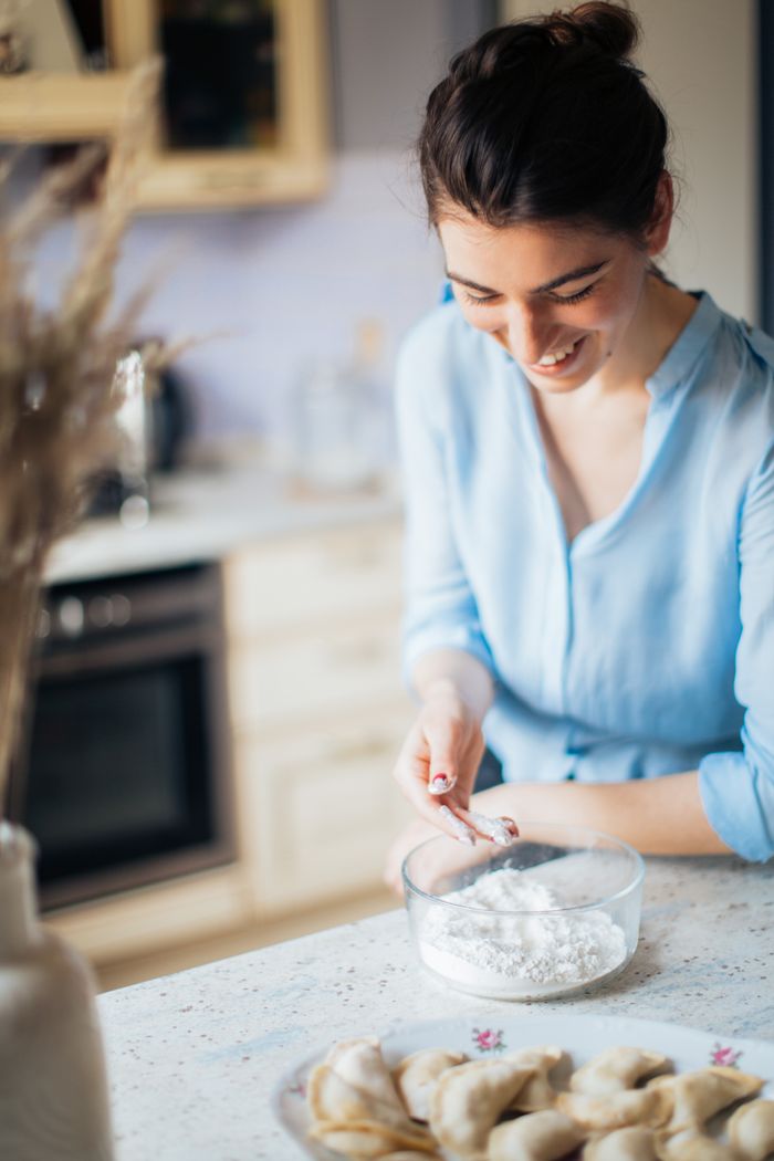 Smiling woman making traditional Russian pelmeni from dough in a cozy kitchen setting.